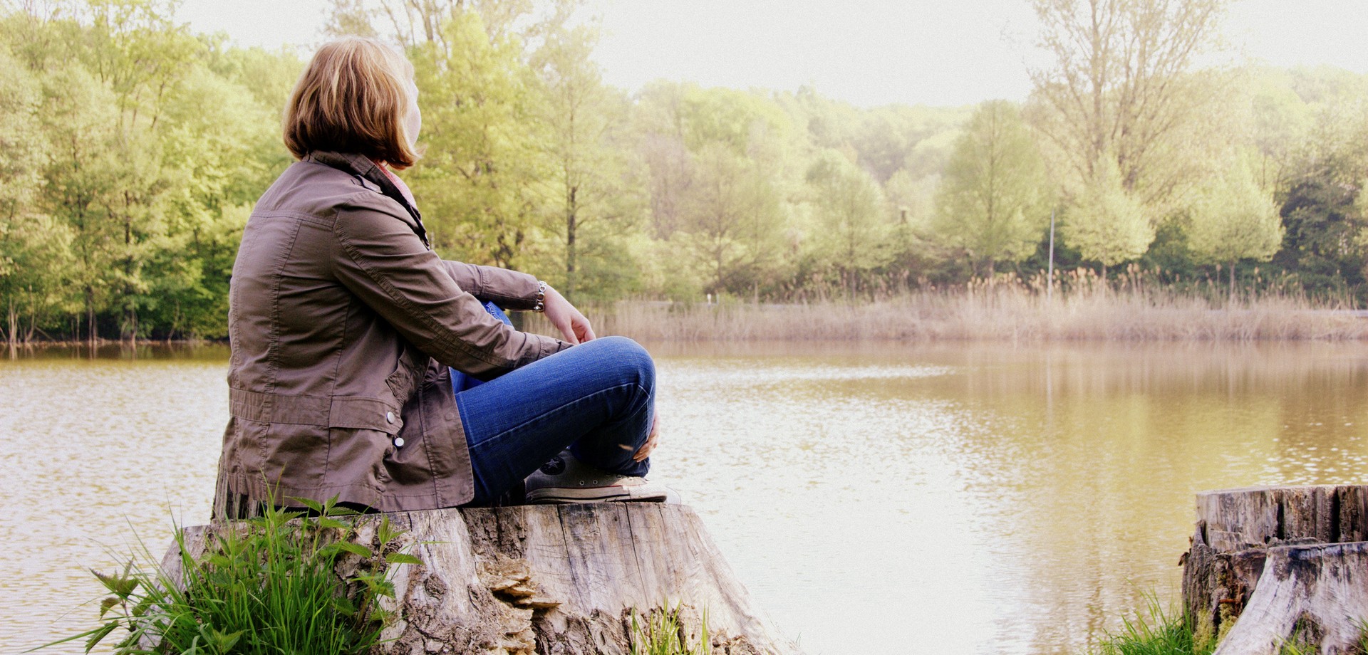 Eine Frau sitzt im Schneidersitz an einem See und blickt auf diesen.